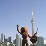 Toy figurine of Indian man in headdress and holding a shield and spear in foreground, urban cityscape with CN Tower in the background