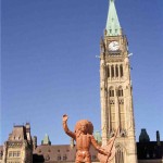 Toy figurine of Indian man in headdress and holding a shield and spear in foreground, Canada's Parliament Buildings and Clocktower in background