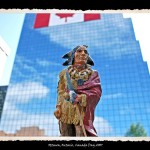 Toy figurine of Indian man in traditional dress and wrapped in red robe in foreground, glass office building with Canadian Flag draped on it in background