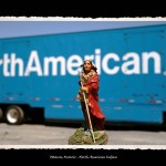 Toy figurine of Indian man in traditional dress and wrapped in red robe in foreground, blue tractor trailer with words North American on it in the background