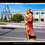 Toy figurine of Indian man in traditional dress and wrapped in red robe in foreground, the glass and concrete structure of the National Gallery of Canada in the background
