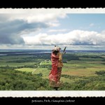 Toy figurine of Indian man in traditional dress and wrapped in red robe on a hill in foreground looking out over grassy vallery and cloudy sky