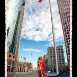 Toy figurine of Indian man in traditional dress and wrapped in red robe in foreground, street scene of tall glass skyscrapers and pole with Canadian flag in background