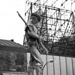 large commemorative statue of man dressed like a cowboy and carrying a shotgun, city buildings behind him