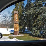 shot from the window of a car of a wooden pole carved with the profiles of two traditionally dressed Indian men, snow, grass and evergreen trees surronding the carving