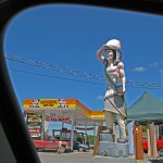 shot framed from a car window, large wooden carving of a statue of an Indian man in traditonal attire and headdress in front of gas station
