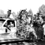 Group of mostly male powwow dancers standing in the back of a pickup truck as it drives away