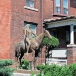 Statue of Indian man in traditional dress on a horse, arms outstretched and face looking upwards, in background brick house in a city neighbourhood