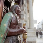 Wooden statue of a cigar store Indian, another one behind him, street scene with people walking and classical architecture with columns in the background