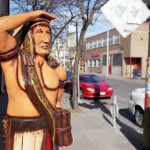 Wooden cigar store Indian standing on urban street scene with parked cars in the background