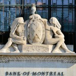Stone carving in front of wrought iron grilles and glass window, carving of two men, one an Indian man in traditional attire, a beaver and an owl stand the crest between them, underneath the words Bank of Montreal are carved