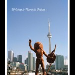 Toy figurine of Indian man in headdress and holding a shield and spear in foreground, urban cityscape with CN Tower in the background